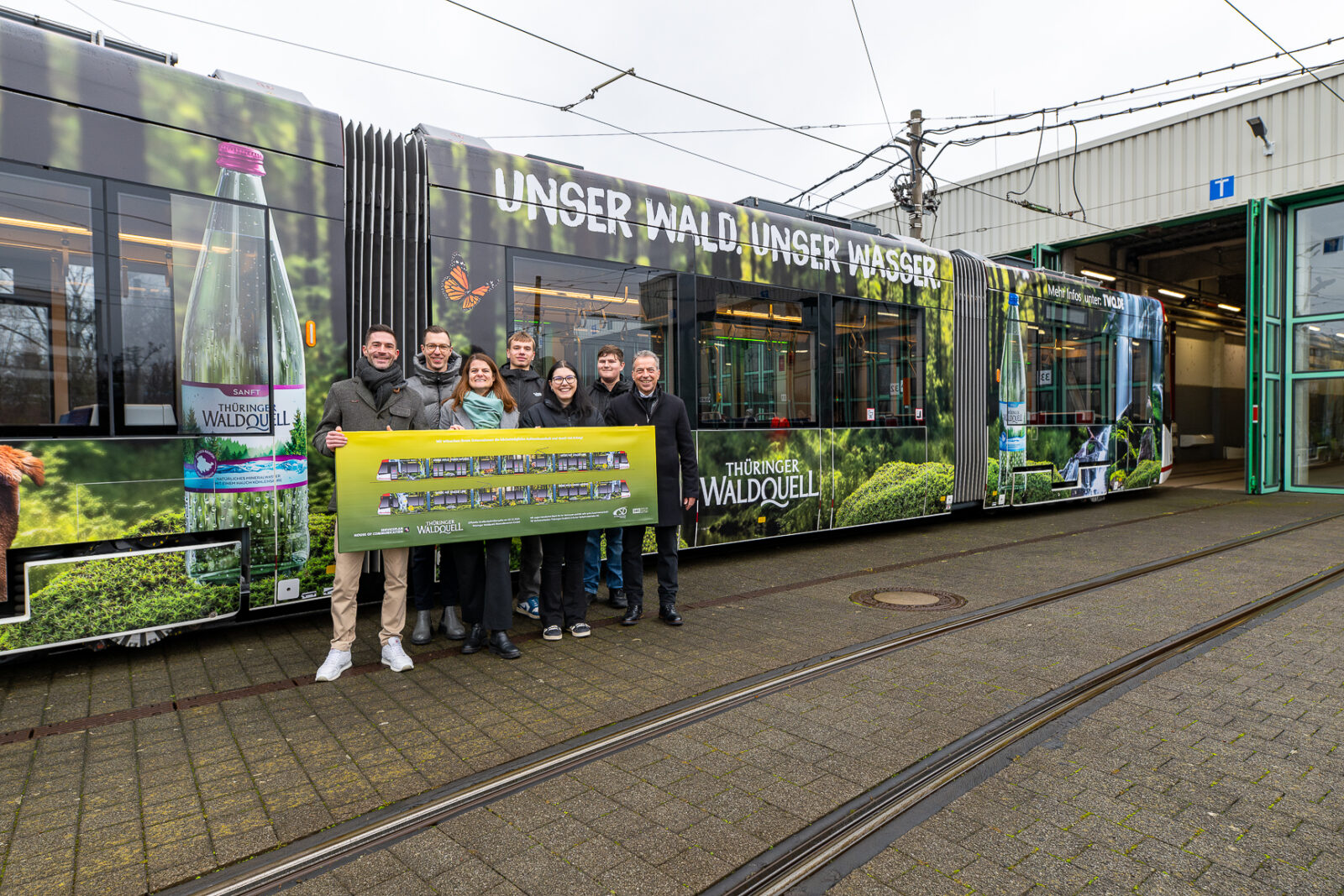 Eine Gruppe von Menschen steht vor einer Straßenbahn und hält ein Bild dieser hoch 