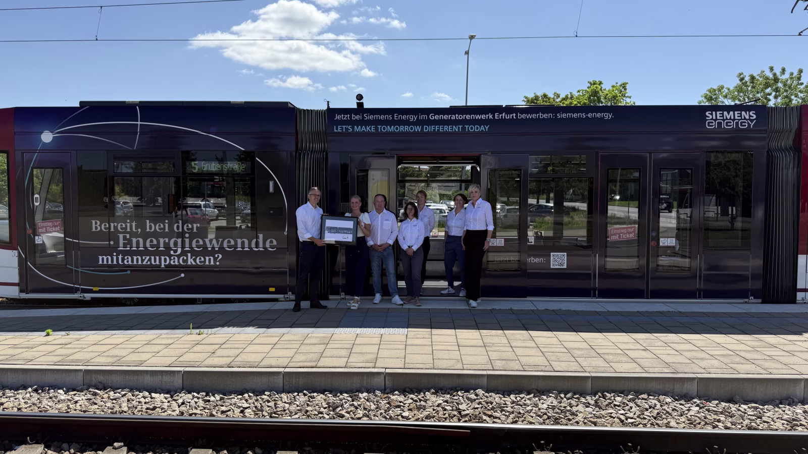 7 Personen stehen vor einer Straßenbahn und halten ein Bild dessen hoch  