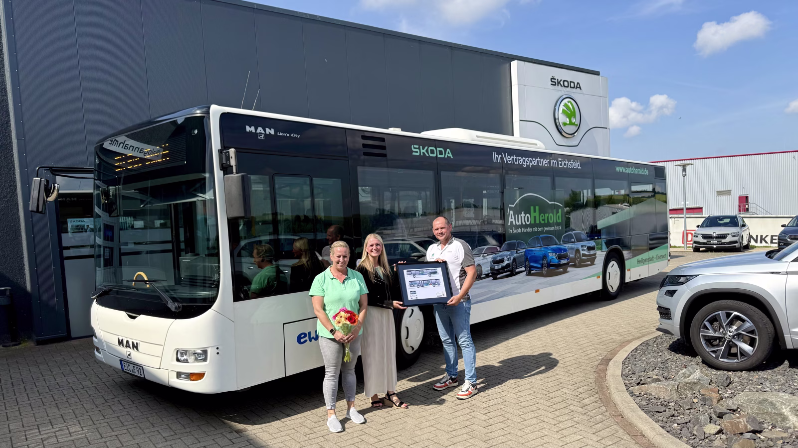Drei Personen stehen vor einem Bus des Autohauses Herold und halten ein Bild dessen in die Luft