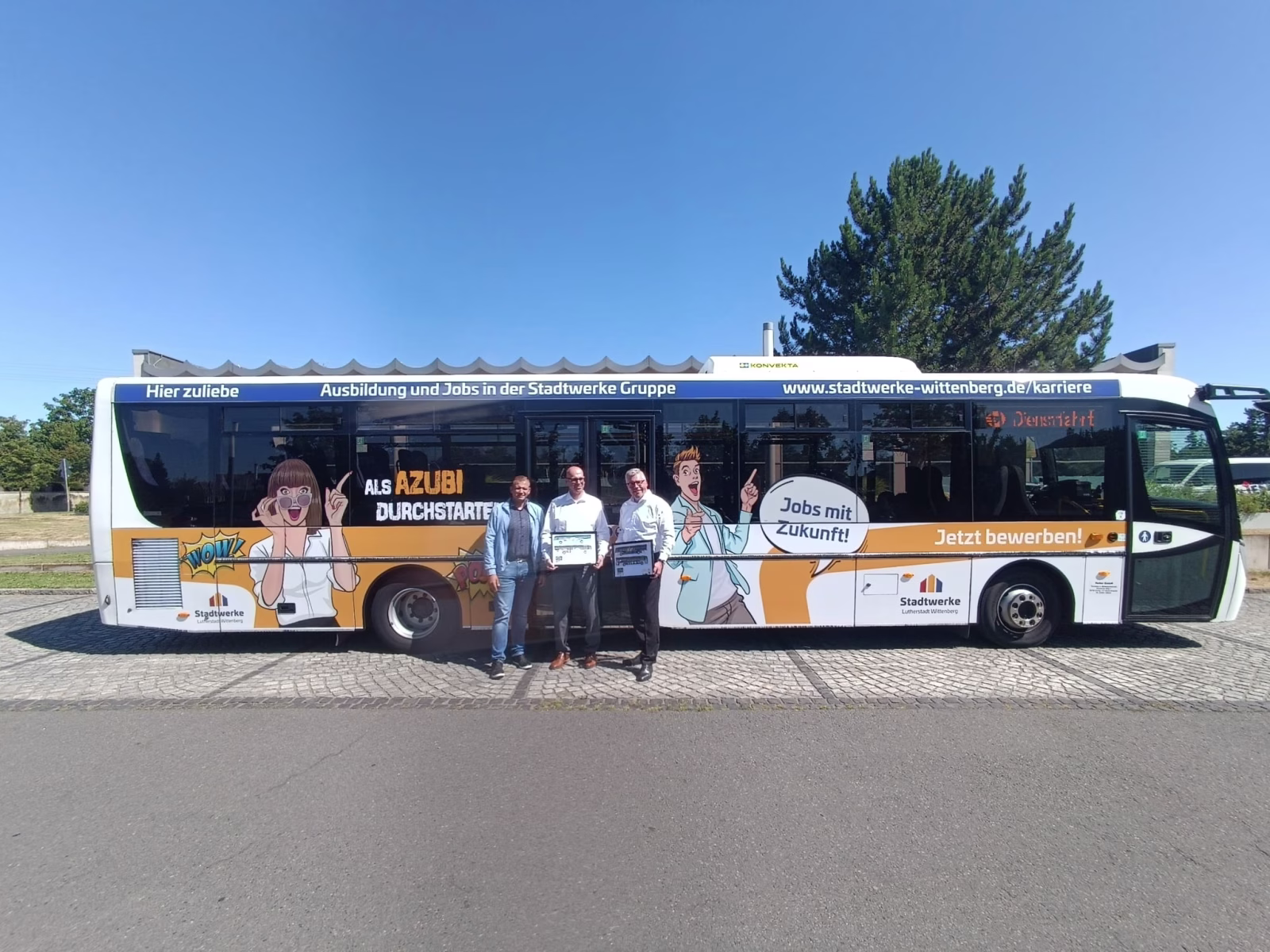 Drei Personen stehen vor einem orangenen Bus und halten ein Bild dessen hoch 