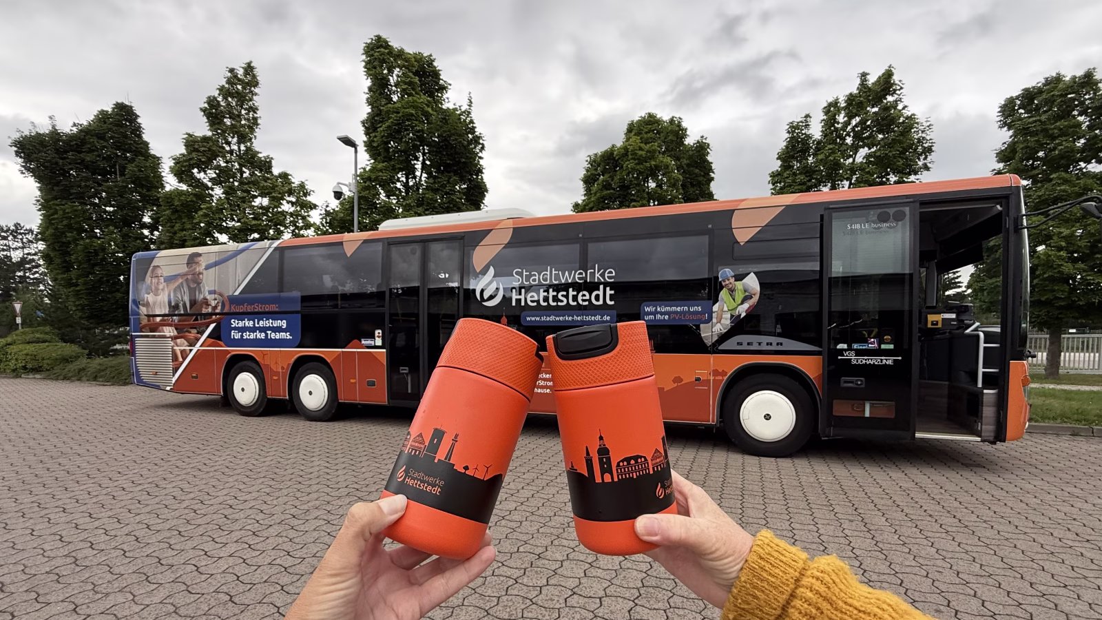 Zwei orange Kaffeebecher werden vor den orangenen Bus der Stadtwerke Hettstedt gehalten 