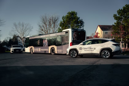 Ein Bus und zwei Autos mit Werbung der Century 21 WohnManufaktur stehen auf einem Parkplatz