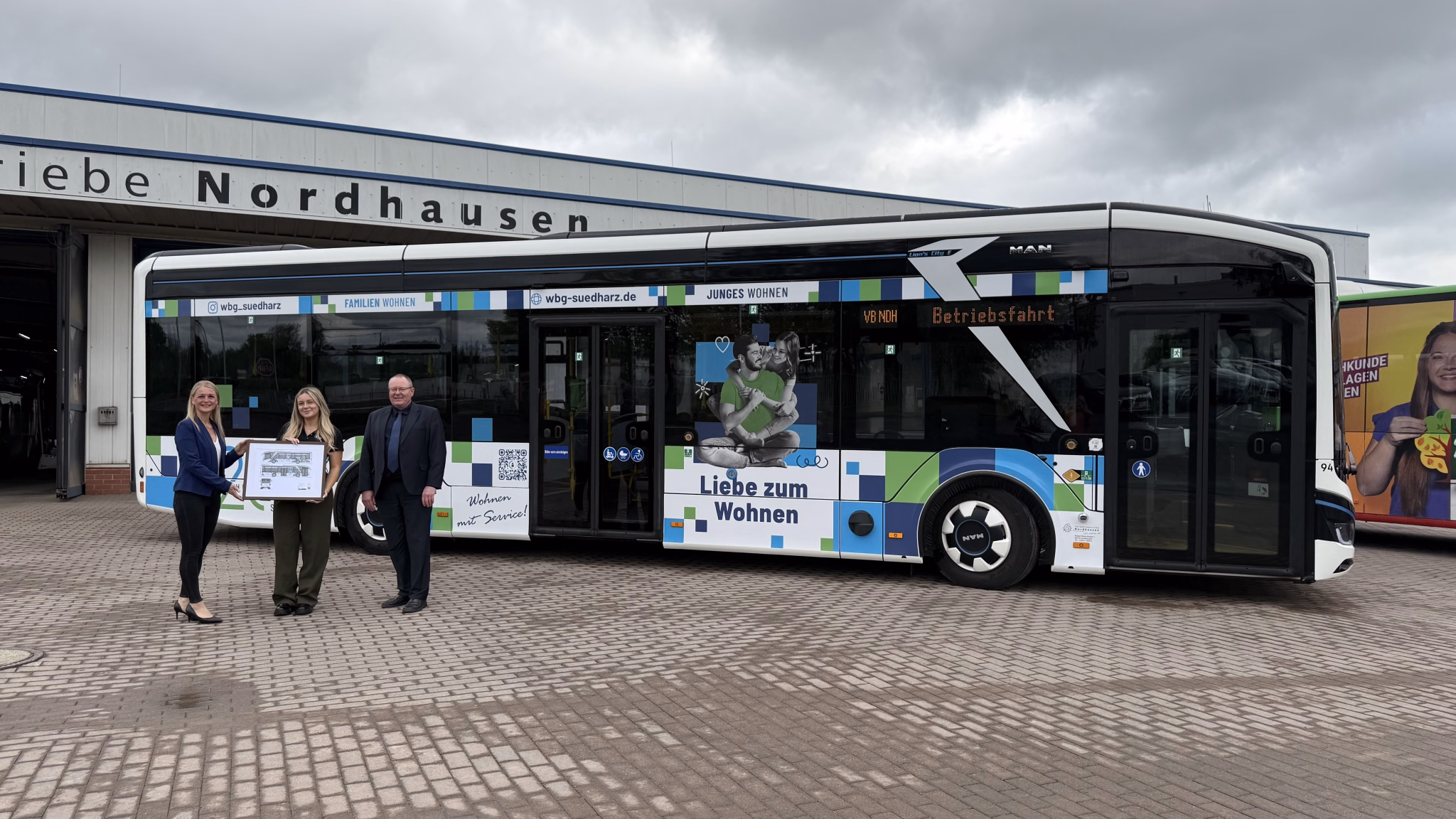 Drei Personen in schicker Kleidung stehen vor einem weißen Bus auf einem Busbetriebshof