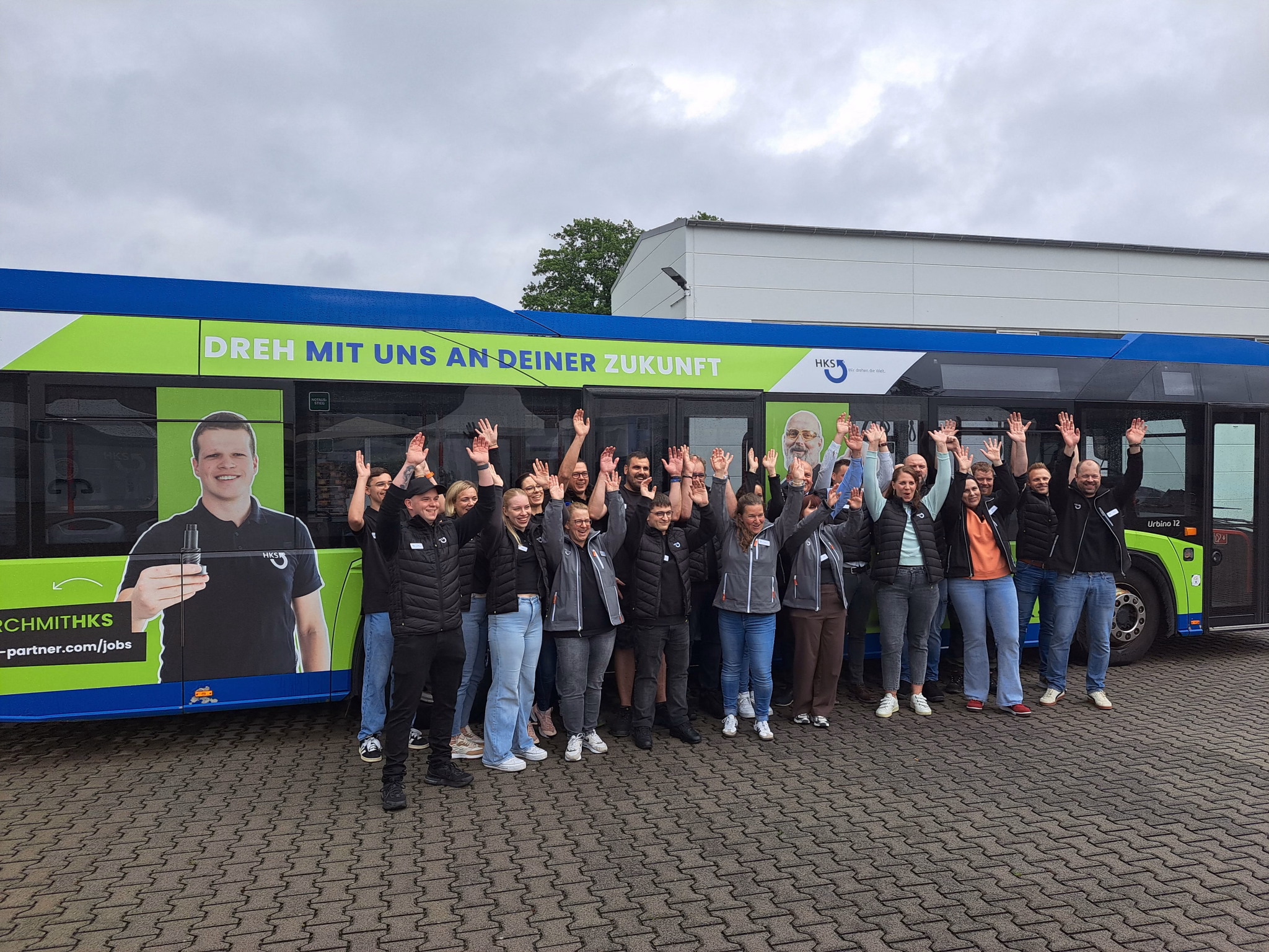 Eine große Gruppe von jubelnden Menschen steht vor einem grünen Bus der HKS Dreh-Antriebe GmbH