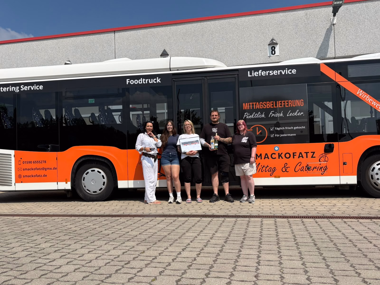 Fünf Personen stehen vor einem orangenen Bus mit Werbung des Smackofatz Catering