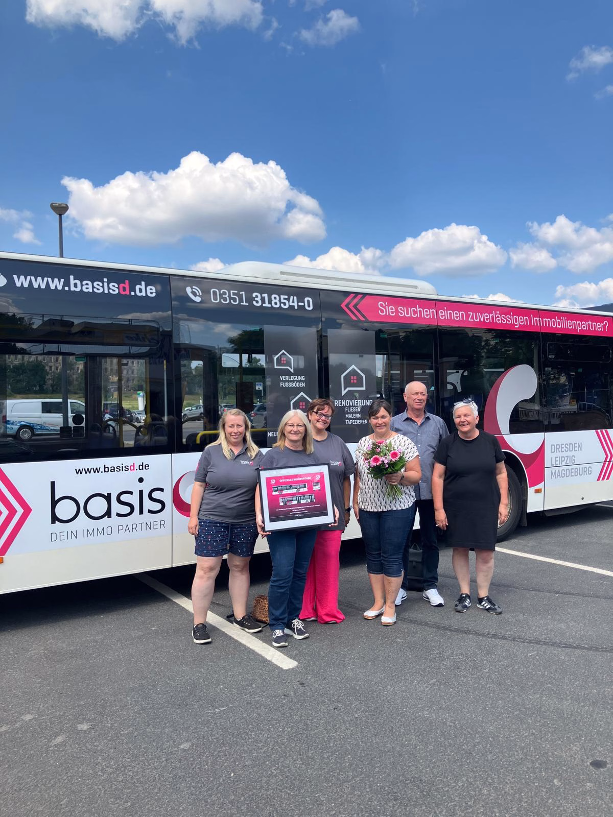 eine Gruppe von Frauen steht vor einem weiß/pinken Bus mit Werbung der Firma basis d