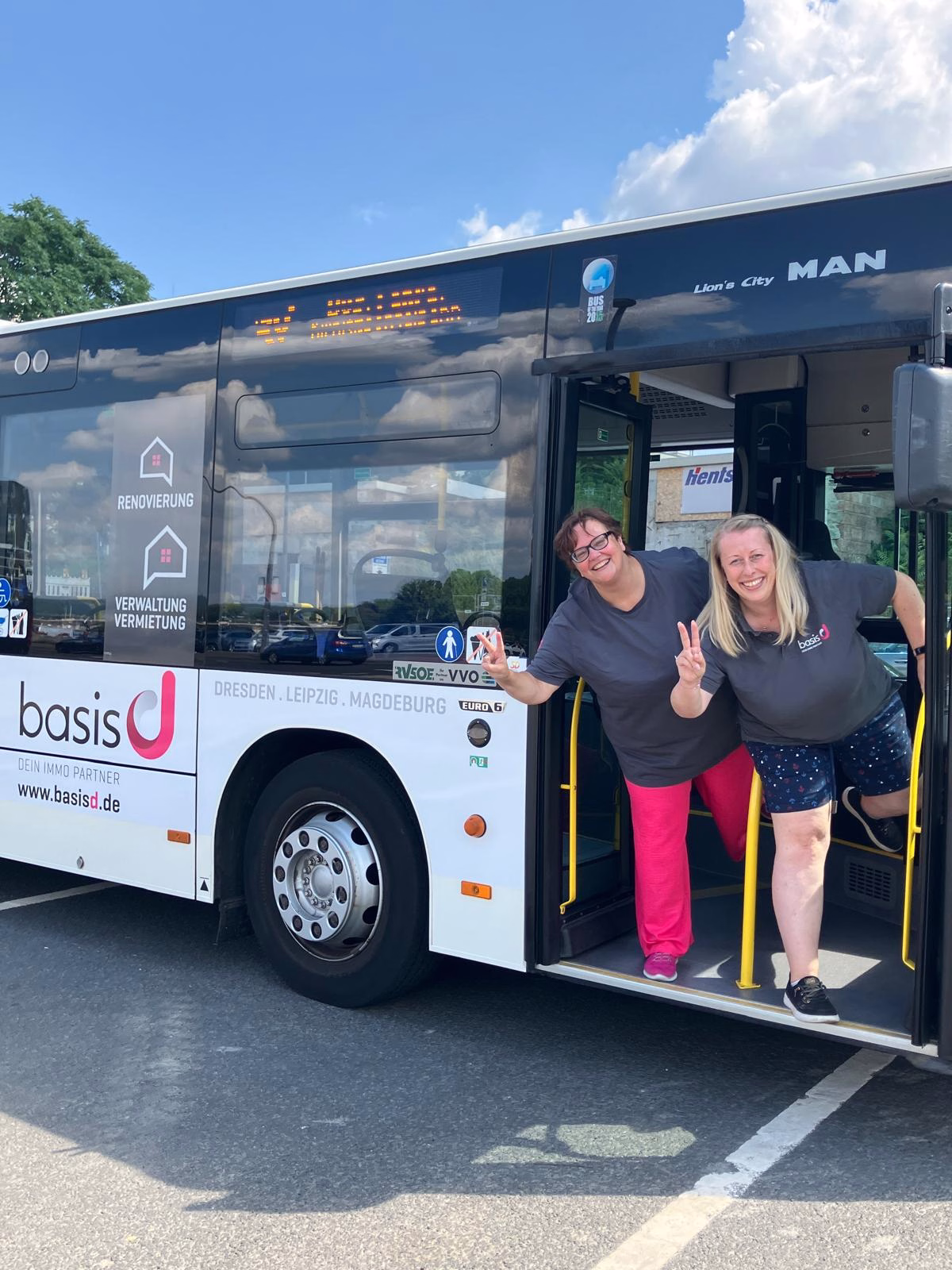 Zwei Frauen winken aus einem Bus mit Werbung des Immobilienbüros basis d