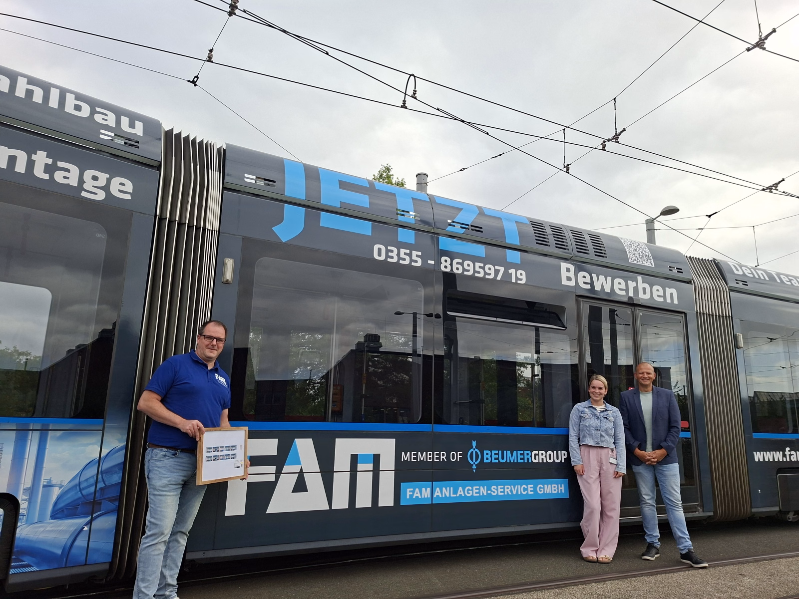 Drei Personen stehen vor einer blauen Straßenbahn mit Werbung der FAM Anlagen