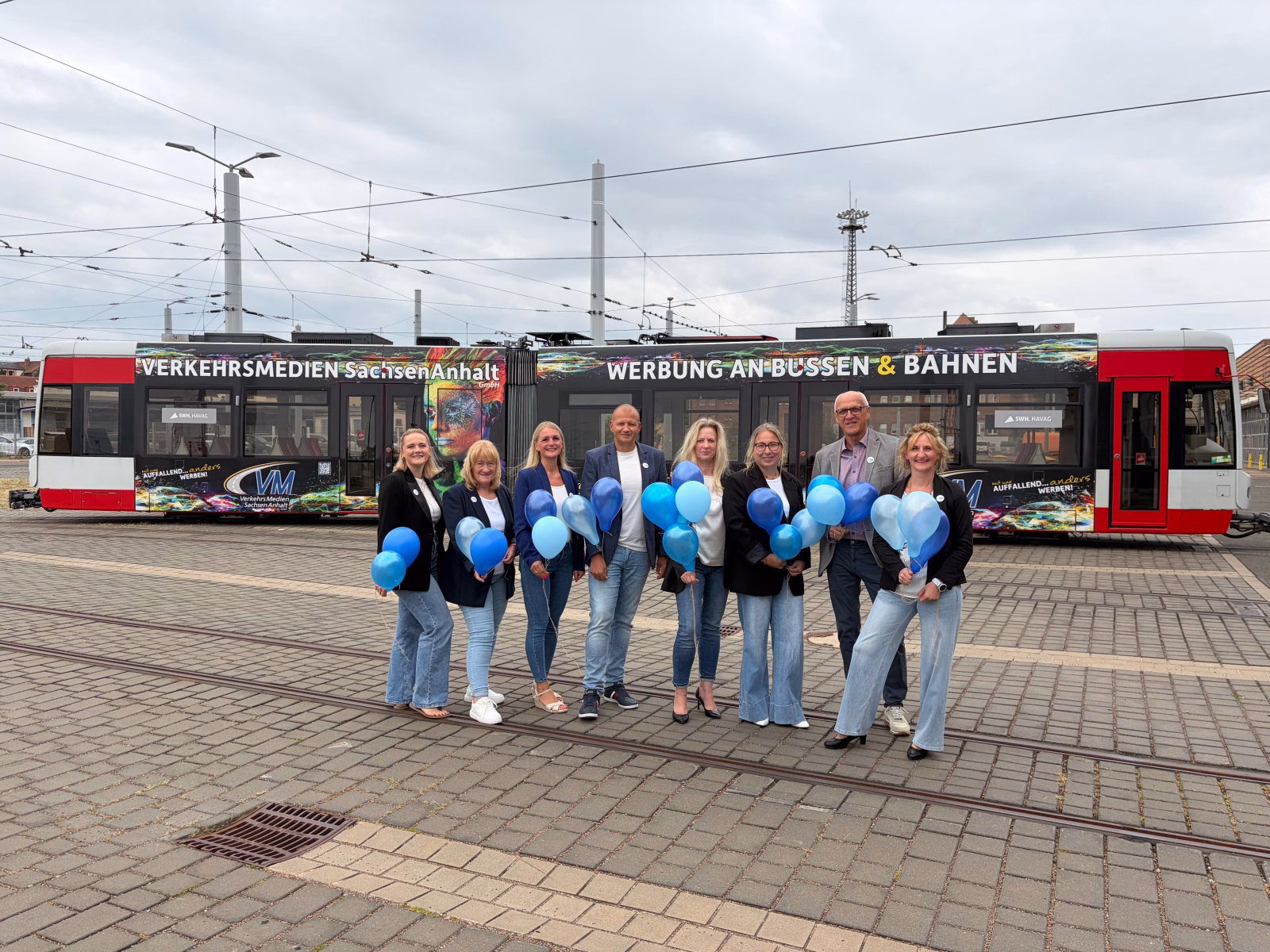 Eine Gruppe von Menschen halten blaue Luftballons hoch und stehen vor einer Straßenbahn
