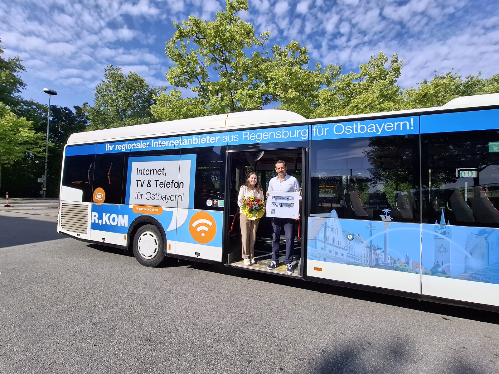 Zwei Personen stehen mit einem Blumenstrauß in der Bustür eines Werbebusses der R-Kom