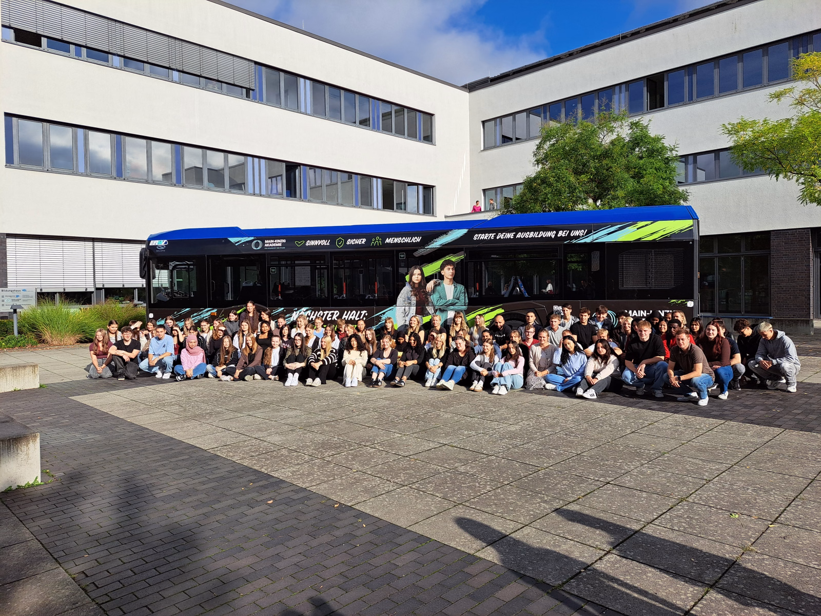 Eine große Gruppe von Azubis sitzen vor einem blauen Werbebus der Main Kinzig Akademie