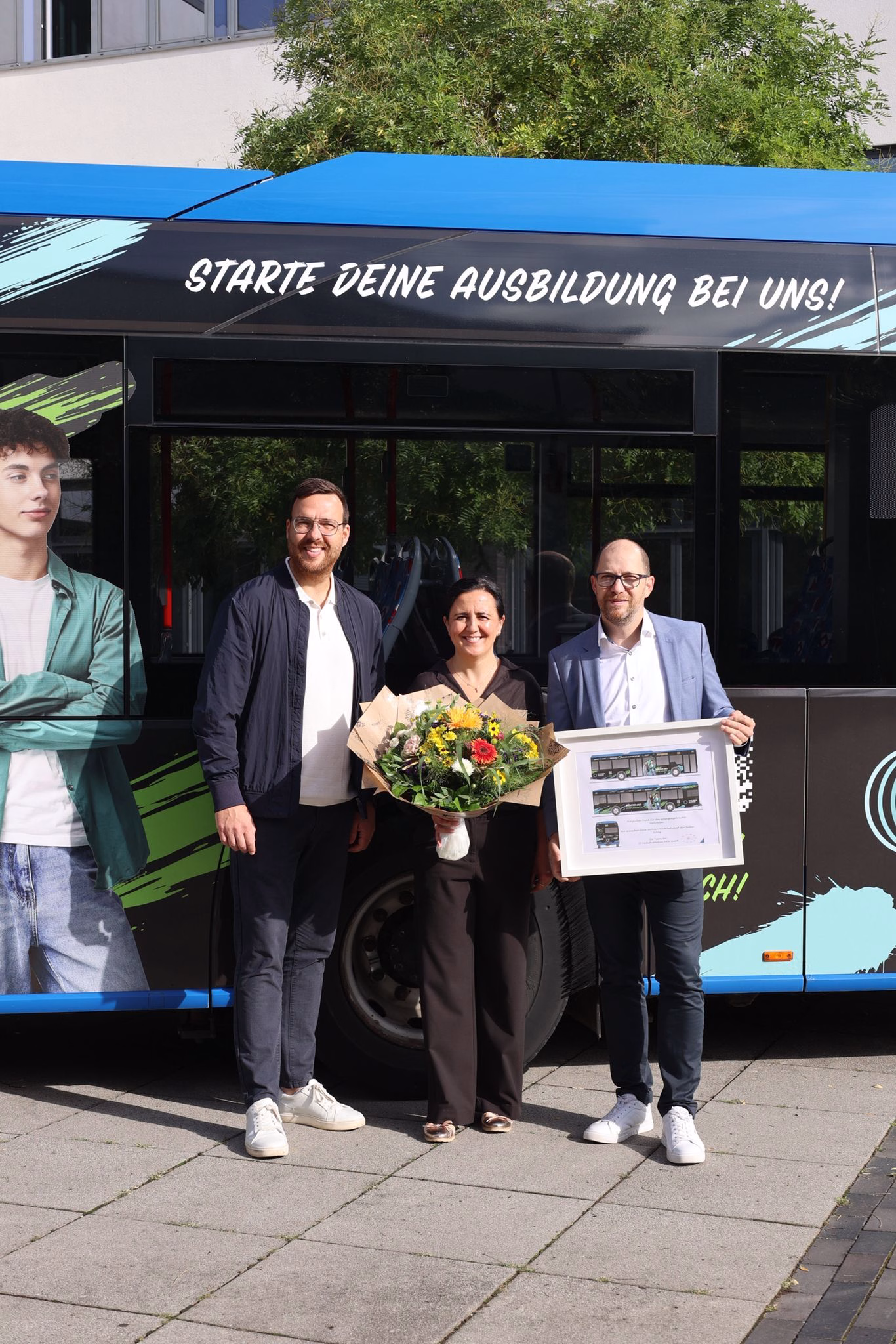 Drei Personen stehen mit einem Blumenstrauß vor einem blauen Werbebus der Main-Kinzig Akademie