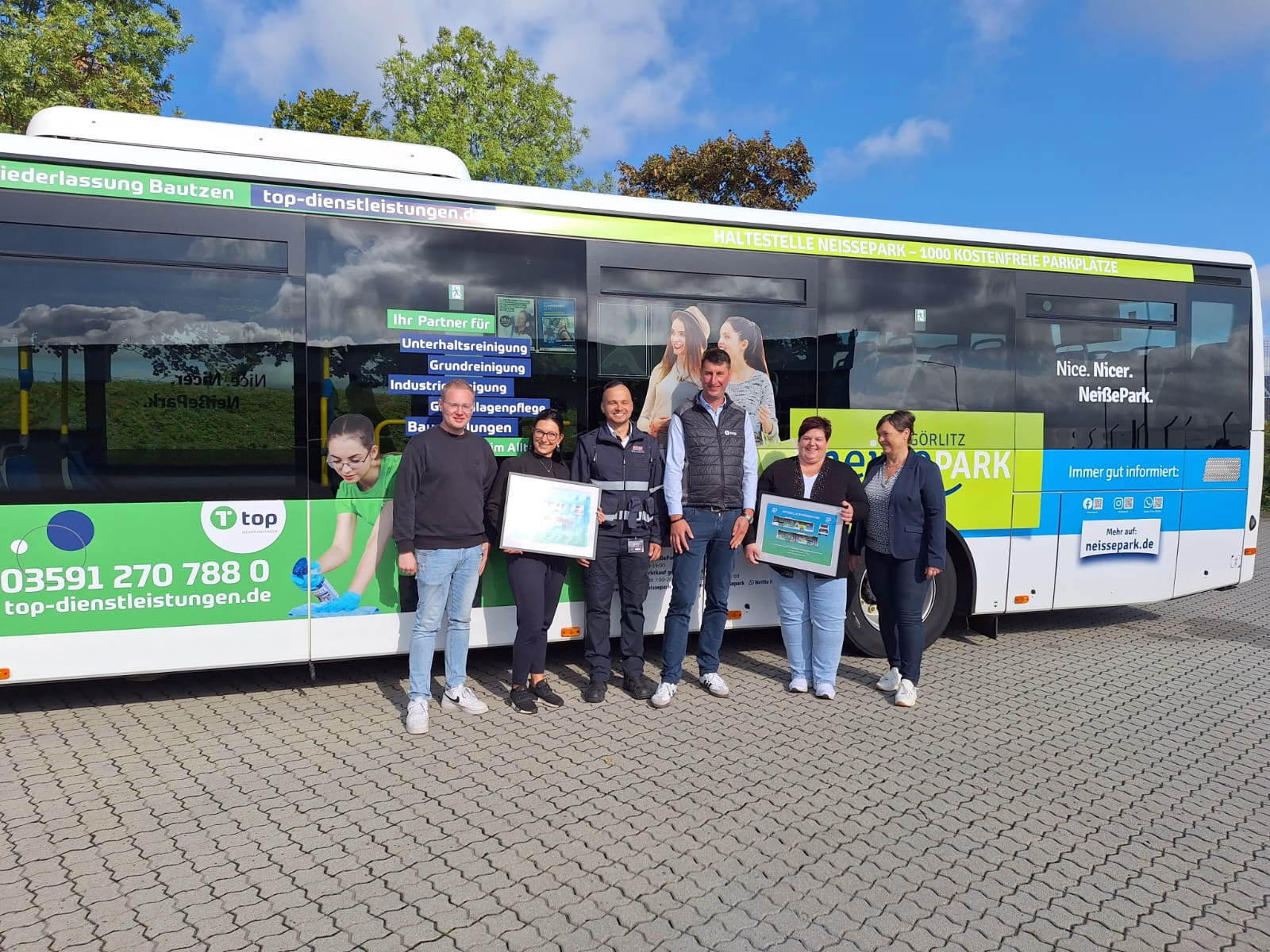 Eine Gruppe von Menschen steht vor einem grün-blauen Stadtbus und lächeln in die Kamera